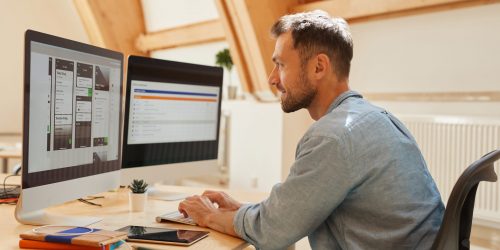 Young programmer sitting at the table typing on computer keyboard and looking at monitor of computer he working at office